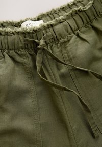 Pantaloni scurți verde măsliniu cu talie elastică, având o margine decorativă cu ciucuri și un șnur legat. Confecționați dintr-un material respirabil.