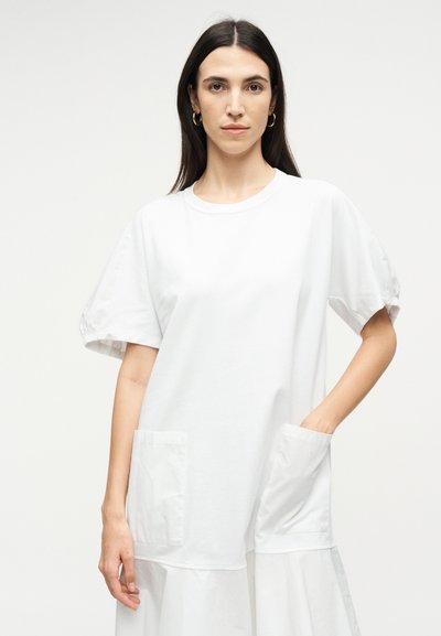 Γυναίκα με μακριά σκούρα μαλλιά, φορώντας ένα άνετο λευκό φόρεμα με τσέπες, με το ένα χέρι στην τσέπη, στέκεται μπροστά σε ένα απλό φωτεινό φόντο.