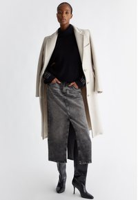 Cappotto beige in lana sopra un maglione nero a maglia e una gonna di denim grigia. Stivali neri al ginocchio con motivo coccodrillo. Taglio verticale sulla gonna.