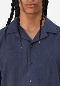 Camisa de cuadros azul marino con botones, confeccionada en tejido texturizado, con un cuello extendido y botones negros. Incluye dos botones en las puntas del cuello.