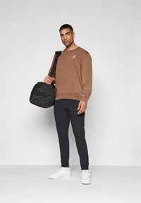 Felpa marrone con logo, pantaloni sportivi neri e sneakers bianche. Porta una borsa a tracolla nera con parte anteriore texturizzata.
