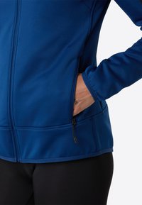 Modrá zipová bunda s hladkým, elastickým materiálem, boční kapsou se zipem a čistým šitím podél švů, nošená přes černé legíny.