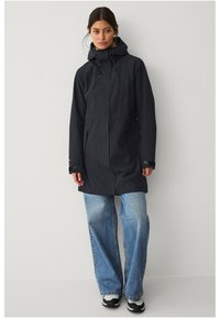 Parka impermeabile grigia scura con cappuccio, caratterizzata da una linea dritta, due tasche laterali e polsini regolabili, abbinata a jeans a gamba larga di un blu chiaro.