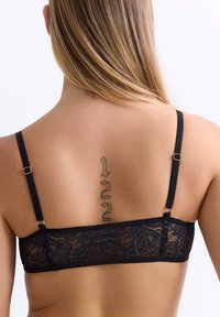Rücken einer Frau mit langen blonden Haaren, die ein vertikales Tattoo zeigt und einen schwarzen Spitzen-BH mit verstellbaren Trägern trägt, vor einem weißen Hintergrund.