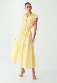 Geltonas, be rankovių suknelė su sagų užsegimu priekyje, raišteliu juosmenyje ir sluoksniuota sijona. Tekstūruotas audinys su atsipalaidavusiu siluetu ir vidutinio ilgio iki blauzdų.