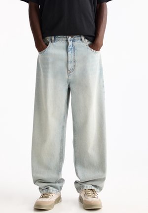 Person trägt locker sitzende, hellblaue Jeans mit Händen in den Taschen und beigen Sneakers, kombiniert mit einem schwarzen Hemd.