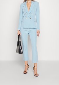 Lichtblauw, op maat gemaakt colbert met dubbele knopenrij en gouden knopen, gecombineerd met bijpassende cropped broek. Houdt een zwarte, getextureerde handtas vast.