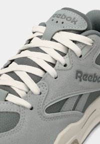 Krupan plan sive Reebok tenisice s bijelim vezicama, mrežastim materijalom, suedenim preklopima i vidljivim logotipom branda na jeziku i središnjem dijelu.