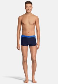 Maillots de bain navy avec une ceinture bleue, présentant un design ajusté et des coutures latérales contrastées, fabriqués à partir d'un matériau lisse et extensible.
