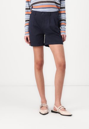 Marintrötta skräddarsydda shorts med veck, gjorda av bomullstyg, matchade med ljusa, lackläderslingbackskor.