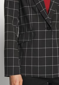 Black and white checkered blazer with notch lapels, a single button, and a side pocket, made from textured fabric.