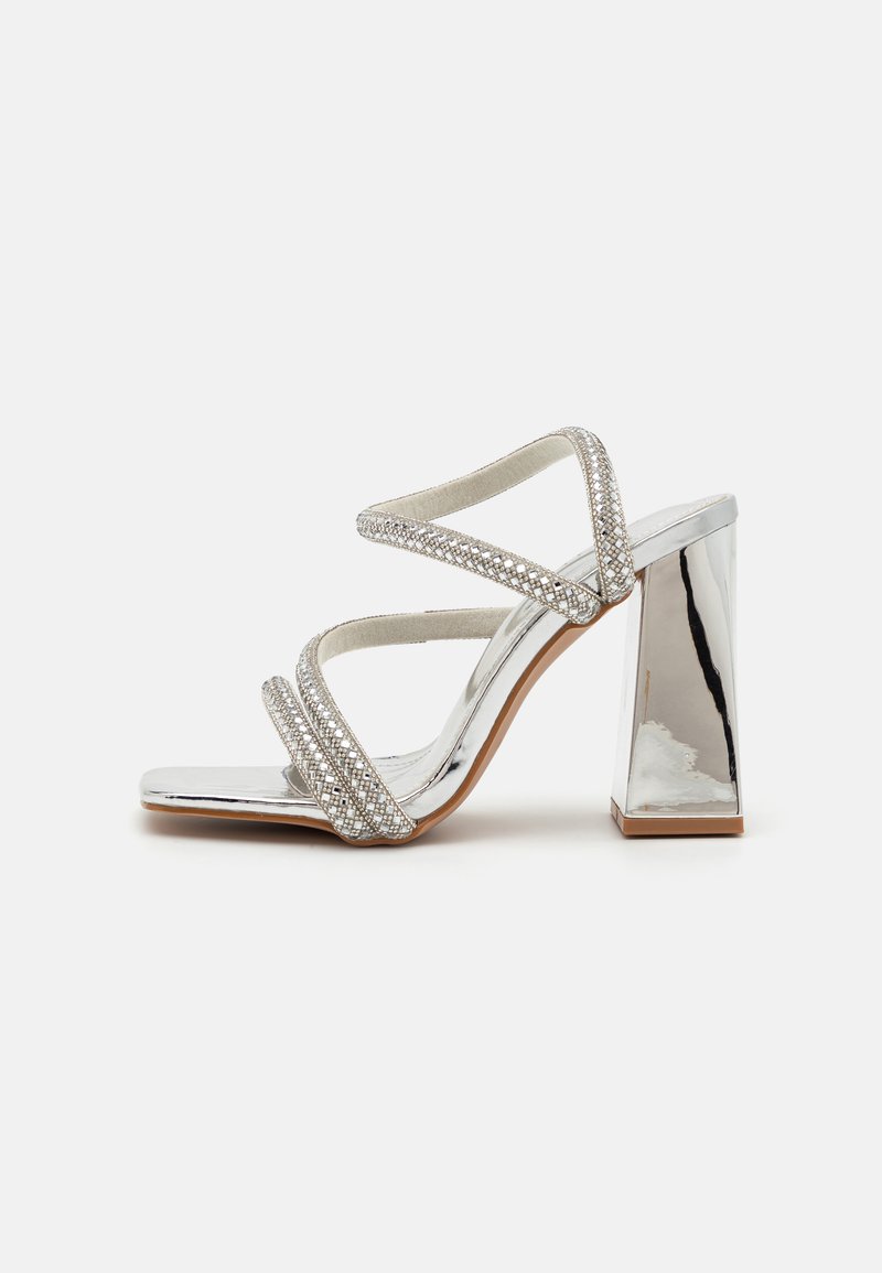 Silver high-heeled sandal featuring cross-strap design, embellished with rhinestones, and a mirrored block heel. Smooth, shiny finish.