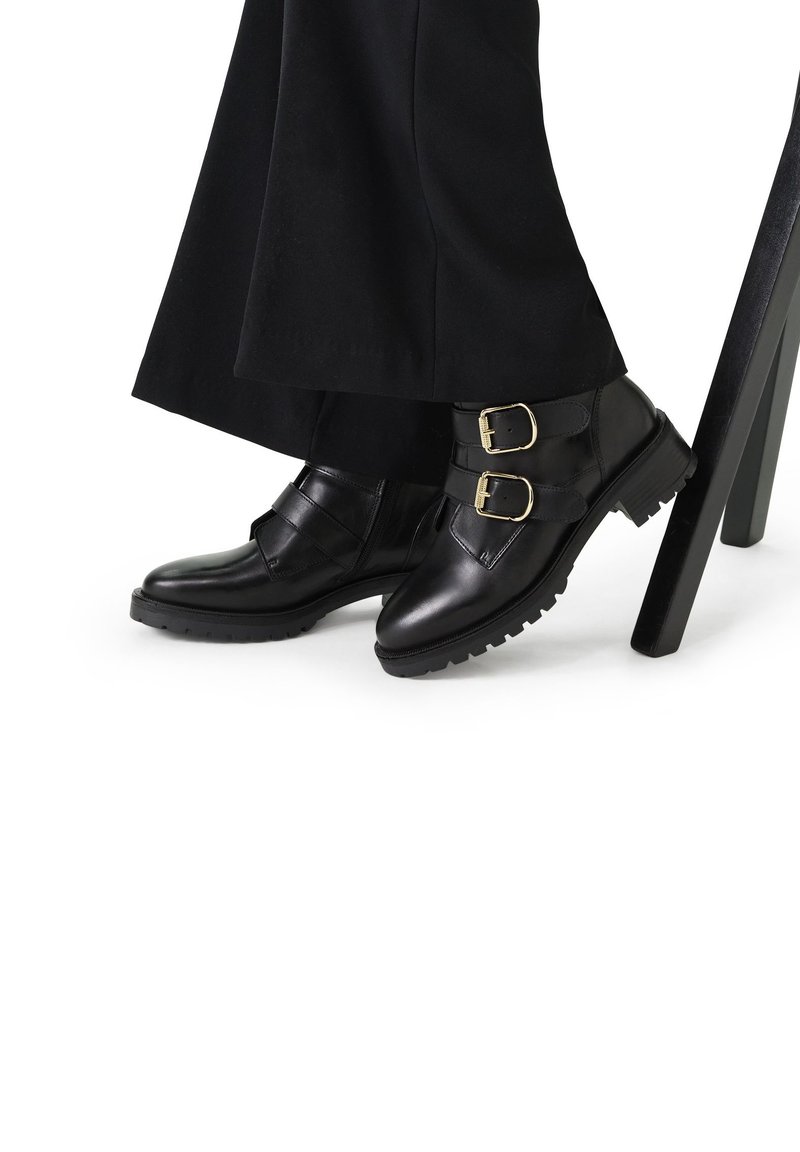 Stivali neri in pelle fino alla caviglia con due fibbie dorate, suole testurizzate e una finitura liscia. Indossati con pantaloni neri lunghi contro il gambo di una sedia.