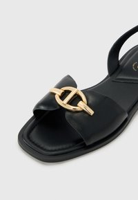 Svart sandal i texturerat läder med en bred rosett och detaljer i guldton. Platt sula med en minimalistisk design.