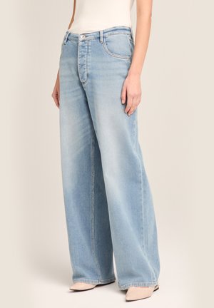 Hellblaue Jeans mit weitem Bein, getragen mit beigen spitzen Schuhen und einem weißen ärmellosen Oberteil, gesehen vom mittleren Oberschenkel abwärts.