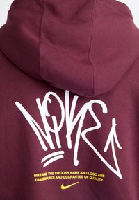 Maroon hoodie featuring a large white graffiti-style logo on the back, with yellow text displaying trademark information. Soft cotton blend material.