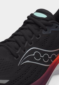 Μαύρο αθλητικό παπούτσι με μαύρα κορδόνια, ασημί λογότυπο της μάρκας στο πλάι, ανοιχτό μπλε ετικέτα στη γλώσσα και σόλα με βαθμιαία απόχρωση από κόκκινο σε λευκό.