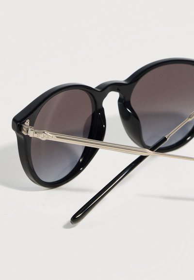 Στρογγυλά μαύρα γυαλιά ηλίου με σκουρόχρωμους φακούς και λεπτούς ασημένιους μεταλλικούς βραχίονες που ακουμπούν σε λευκή επιφάνεια.