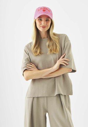 Jeune femme aux longs cheveux blonds portant une chemise beige oversize et un pantalon, ainsi qu'une casquette rose avec l'inscription "AMORE" et un cœur.