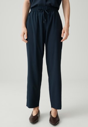 Person trägt marineblaue Hose mit Kordelzug und dunkelbraune spitze Schuhe, steht vor einem einfarbigen hellen Hintergrund.