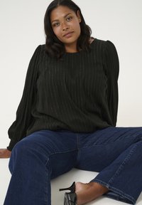 Blouse noire avec des rayures verticales en argent, manches bouffantes, portée avec un jean évasé bleu et des chaussures à talons hauts noires.