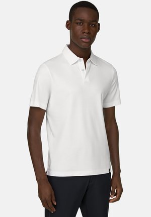 Młody mężczyzna o ciemnej karnacji, ubrany w prostą białą koszulkę polo z krótkim rękawem i czarne spodnie, stojący na jasnym tle.