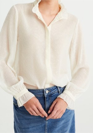 Bílá texturovaná halenka s volánkovým límcem a manžetami, s knoflíkovou zapínáním vpředu, kombinovaná s džínovou sukní. Lehká, vzdušná látka.