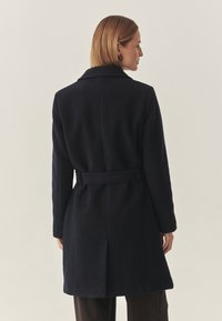 Marineblauer Wollmantel mit tailliertem Gürtel, Reverskragen und langen Ärmeln. Besitzt eine glatte Textur und einen geraden Schnitt, der bis zur Mitte des Oberschenkels reicht.