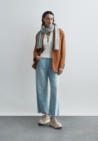 Sacou din catifea maro, top alb din tricot cu detalii cu șnur, blugi largi de culoare albastră deschisă, eșarfă gri și adidași platformă bej.