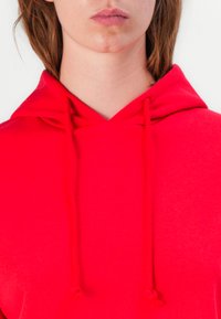 Sarkans kapuci džemperis ar aukliņu, izgatavots no mīksta auduma. Dizains ir vienkāršs, ar brīvu piegriezumu un minimālu detaļu skaitu.