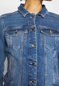 Close-up of person wearing a blue denim jacket with metal buttons and two chest flap pockets, collar visible, head partially shown.