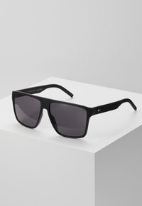 Lunettes de soleil noires rectangulaires avec des verres foncés. Monture noire mate avec des côtés texturés et un petit logo sur la branche. Design enveloppant de Joan.