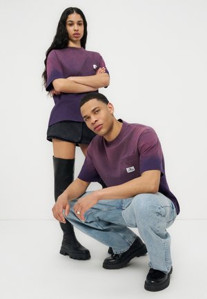 Frau steht mit verschränkten Armen in einem lila Farbverlauf-Shirt, schwarzem Rock und Stiefeln; Mann hockt in passendem Shirt und hellen Jeans.