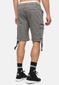 Shorts cargo gris avec une taille élastique à cordon, plusieurs poches et des zips sur les côtés. Associés à des chaussures de sport noires et des chaussettes à rayures.