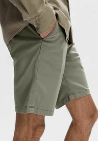 Pantaloni scurți din bumbac verde, cu un croi relaxat. Au buzunare laterale și margini răsucite. Purtați cu o cămașă deschisă khaki cu nasturi.