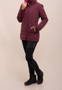 Veste isolée bordeaux avec capuche zippée, poches avant et accents noirs, associée à un pantalon noir ajusté et des bottes de randonnée noires.