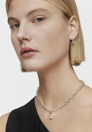 Collana in argento sterling con un design massiccio e un piccolo pendente che assomiglia a un orsacchiotto, indossata con una maglietta nera.