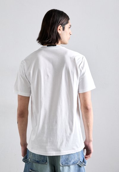 Camiseta de algodón blanca de manga corta y escote redondo, con una etiqueta de logo en la parte de atrás, combinada con pantalones cortos de mezclilla azul claro.