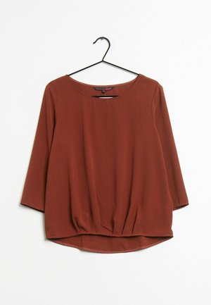 Blusa de mujer de manga larga color óxido, con escote redondo, corte holgado y ligero detalle fruncido en la parte inferior, colgada en una percha negra.