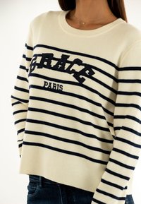 Maglione bianco con righe orizzontali blu navy, con la scritta "GRACE" e "PARIS" in bold. Tessuto morbido, colletto rotondo, vestibilità leggermente rilassata.