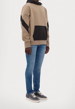 Person wearing beige and black hoodie, blue slim-fit jeans, and gray sneakers standing against a white background.