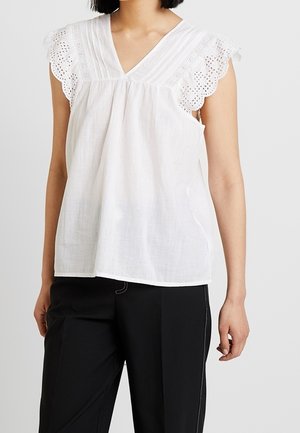 Nainen pukeutuneena valkoiseen hihattomaan puseroon, jossa on silmukkapitsi hihoissa, ja mustiin housuihin, seisoo yksiväristä taustaa vasten.