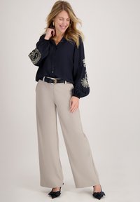 Marineblouse met geborduurde manchetten, knoopsluiting aan de voorkant en wijd uitlopende mouwen, gecombineerd met beige broek met wijde pijpen; zwarte riem en hakken met strikversiering.