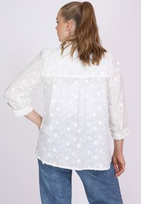 Weiße bestickte Bluse mit floralem Muster, durchscheinendem Stoff, langen Ärmeln mit Rüschensaum und lockerer Passform, kombiniert mit Jeanshosen.