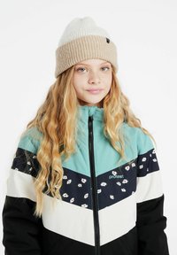 Enfant portant une veste multicolore avec une fermeture éclair, ornée de motifs floraux, en bleu-vert, noir et blanc. Bonnet en tricot beige et crème.