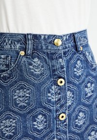 Denim skirt with embossed floral patterns and gold buttons, worn with a tucked white top.