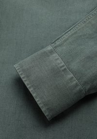 Puño de camisa de lino verde con una textura suave y detalles de costura visibles. La tela parece ligera y transpirable.