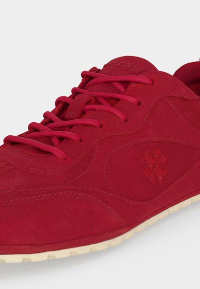 Gros plan d'une basket en suède rouge avec des lacets ton sur ton, un logo embossé sur le côté et une semelle en caoutchouc blanche visible sur le bord inférieur.