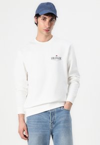 Hvit genser med lange ermer, rund halslinje og et lite rødt og blått emblem på brystet. Kombinert med lyseblå denimjeans og en marineblå caps.