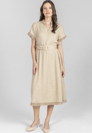 Kobieta w beżowej sukience midi z frędzlami na rękawach i dole, z paskiem podkreślającym talię, stojąca na tle jasnej, gładkiej ściany.
